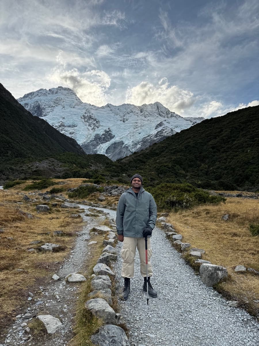 Prathap standing on a mountain trail in New Zealand, snow-capped peaks behind — founder of Men-Kind